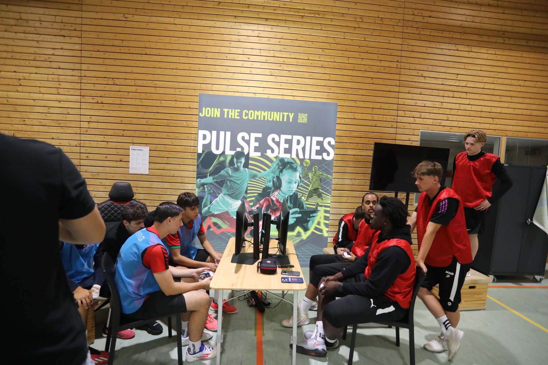 PULSE Series matchday in a sports hall — consoles and court in one venue Darkened gym during a PULSE Series matchday: players at the consoles in the foreground, team-mates on the court behind them
